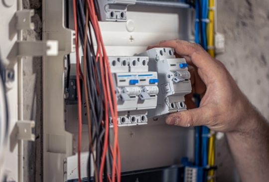 A male electrician works in a switchboard with an electrical connecting cable, connects the equipment with tools.